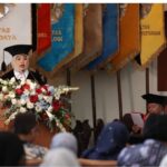 Female graduate in cap and gown speaks at the podium with a large floral arrangement colorful banners behind her